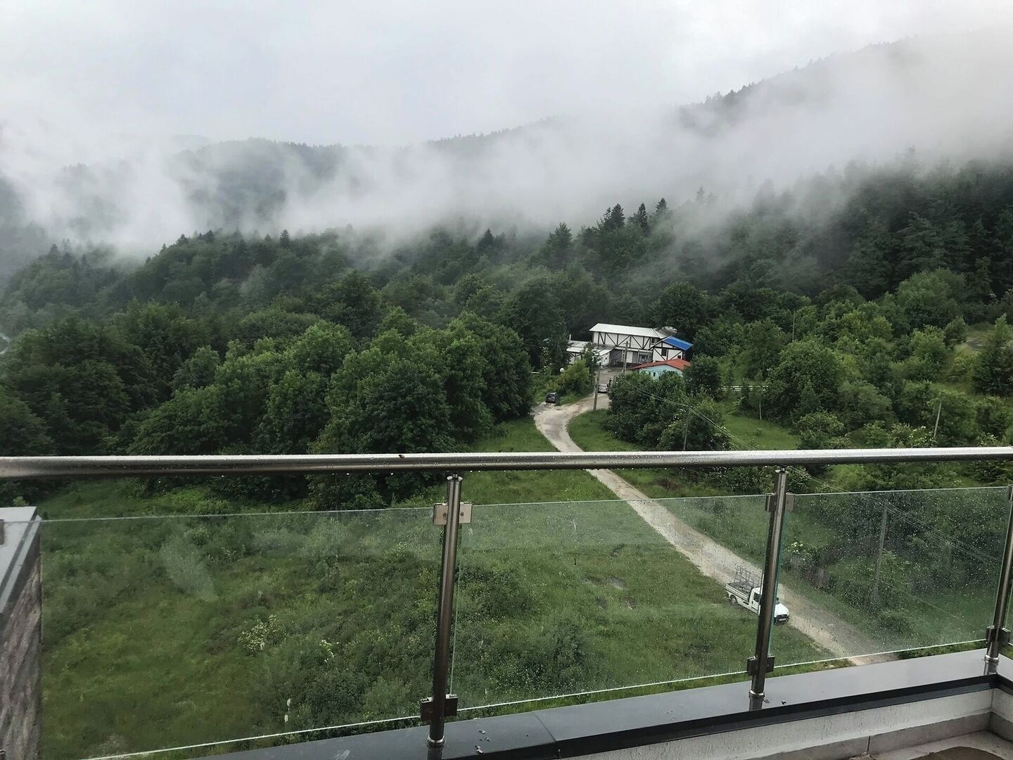 A view from a balcony overlooking a forested valley with a road leading to a building with a red roof.