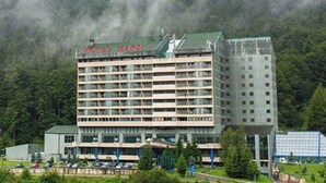 A multi-story hotel with a green roof, surrounded by lush greenery and a forested backdrop.