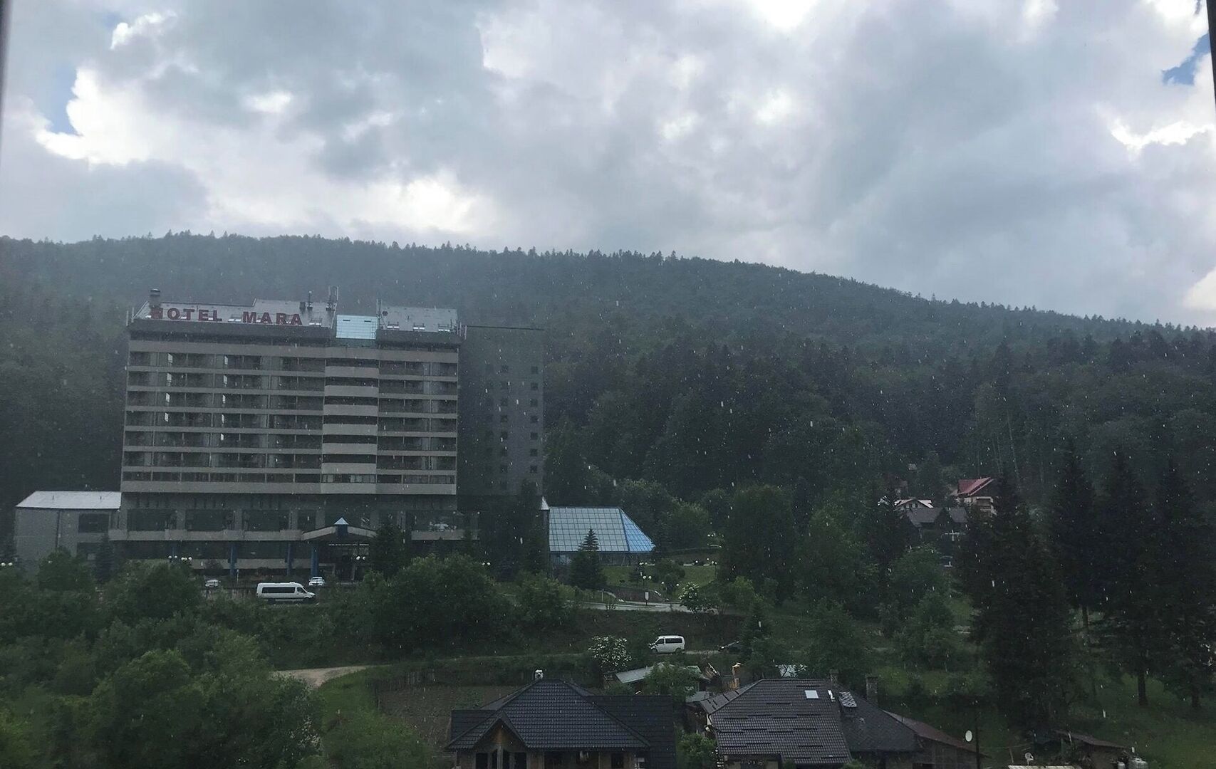 A multi-story hotel with a sign reading 'HOTEL MARA,' surrounded by greenery and a mountain in the background.