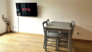 Ett rum med ett matbord och stolar, en väggmonterad tv som visar ”NETFLIX”, och en vas med blommor på golvet.