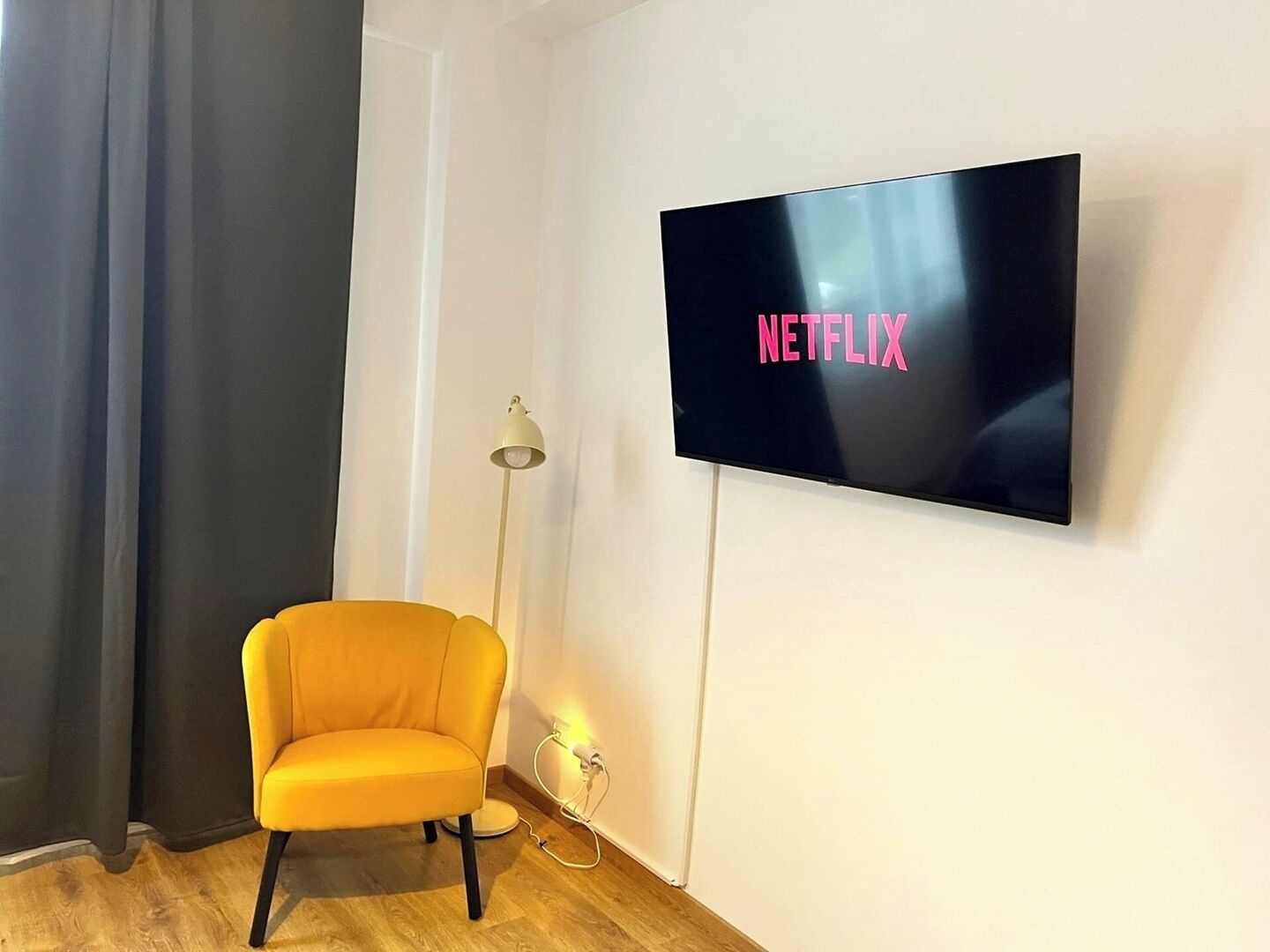 Een kamer met een wandgemonteerde flatscreen televisie waarop ‘NETFLIX’ in roze letters wordt weergegeven, een gele fauteuil, een vloerlamp en een stopcontact.