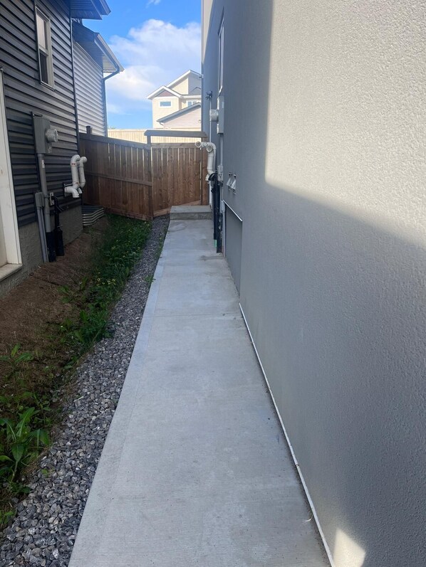 A narrow concrete pathway leading to a wooden fence, with a grey building on one side and a white building on the other.