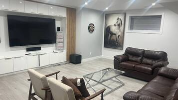 A modern living room with a large flat-screen TV, leather sofas, a glass coffee table, and a dartboard on the wall.