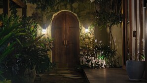 Une porte en bois surmontée d’un arc en pierre, entourée d’une végétation tropicale luxuriante et donnant sur une terrasse en bois qui mène à un grand pot de fleurs.