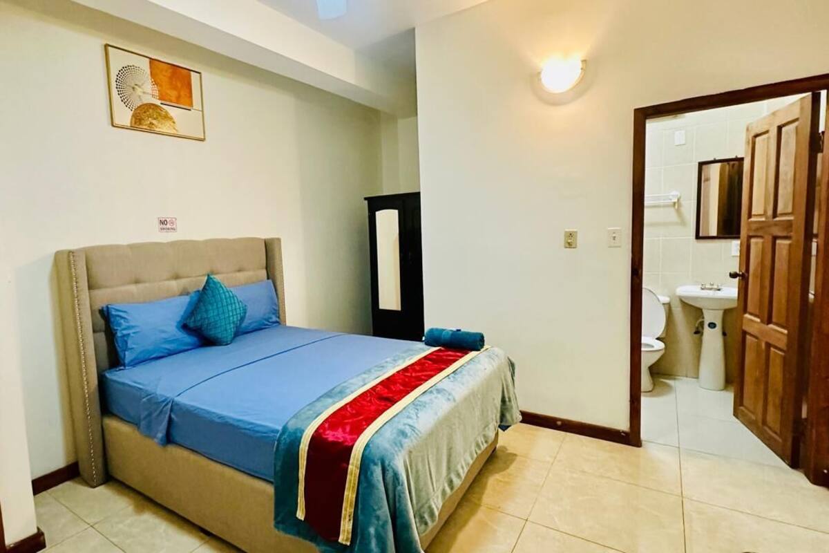 A bedroom with a bed, a bedside table, a mirror, and a bathroom visible through an open door.