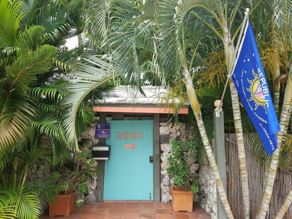 L’entrée d’un immeuble est marquée par une porte turquoise portant le numéro « 1001 », et est entourée de plantes tropicales et d’un drapeau bleu orné d’un symbole solaire.