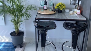 Una cocina moderna con encimera de mármol, dos taburetes negros, una planta en maceta y una estantería para vinos con botellas.