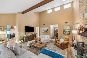 A spacious living room with a fireplace, a television, and a dining area with a kitchen in the background.