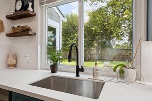 A kitchen with a stainless steel sink, a potted plant, a bottle of soap, and a window with a view of a backyard.