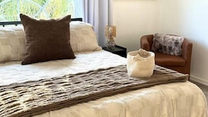 Une chambre avec un grand lit, une fenêtre avec des rideaux, un tableau accroché au mur, une table de chevet et une chaise.