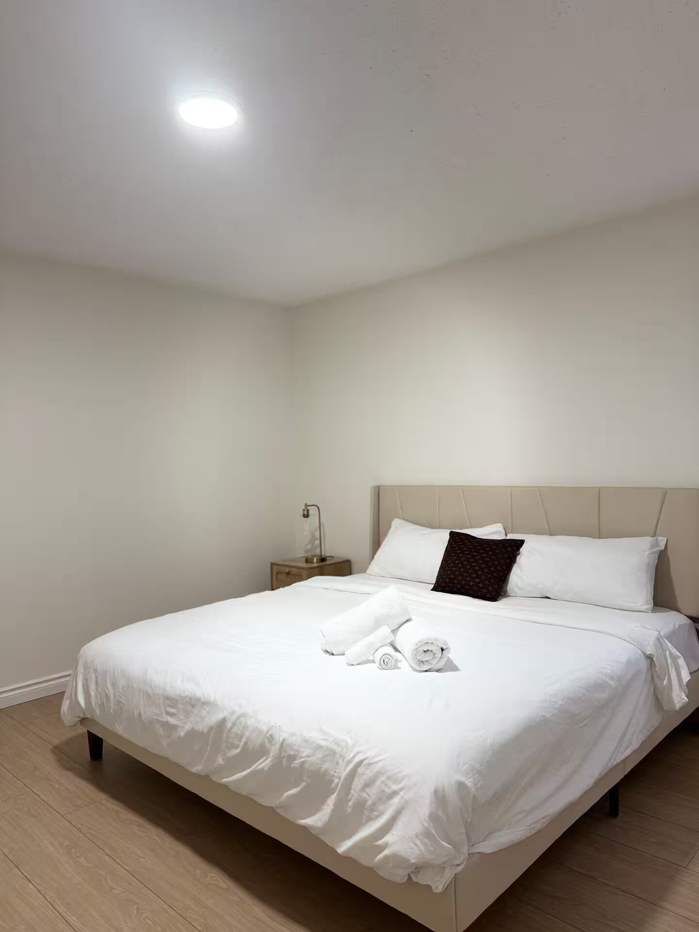 Una cama bien hecha con sábanas blancas y una toalla enrollada, una mesita de noche con una lámpara y una pared blanca lisa.