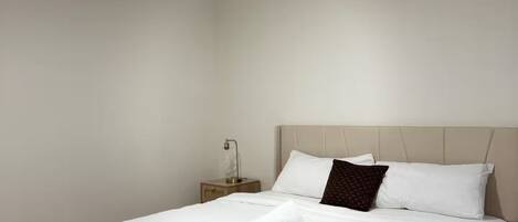 Una cama bien hecha con sábanas blancas y una toalla enrollada, una mesita de noche con una lámpara y una pared blanca lisa.
