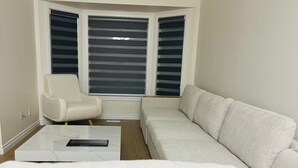 Een hoekappartement met een witte bank, een witte fauteuil, een glazen salontafel en een erker met zonwering.