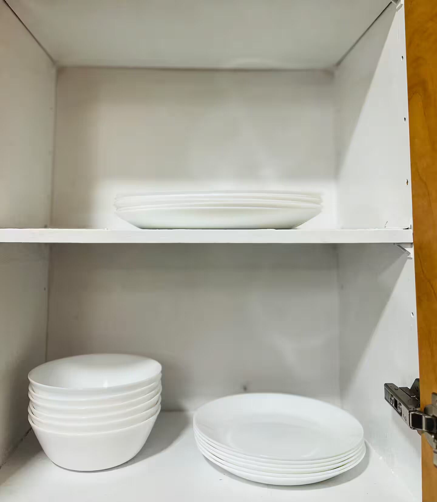 A white cabinet with two shelves; the top shelf holds a stack of white plates, and the bottom shelf has a stack of white bowls.