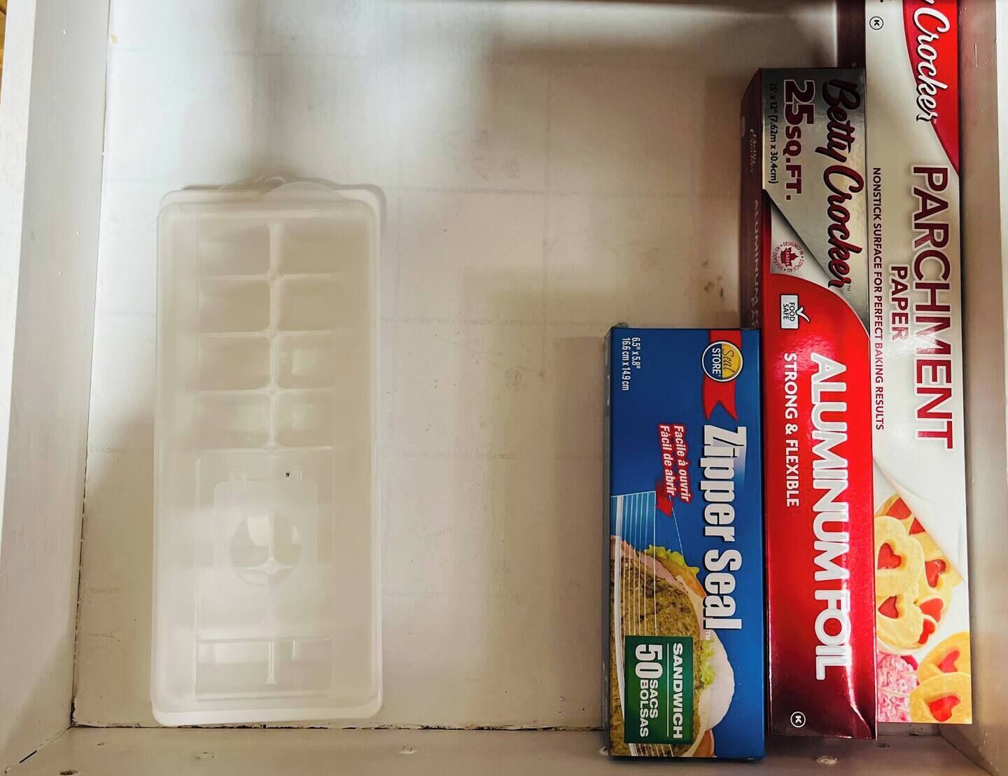 A kitchen shelf with a plastic container, a box of Zipper Seal, a box of aluminum foil, and a box of parchment paper.