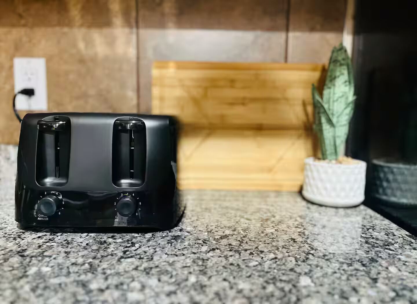 A black toaster on a granite countertop with a wooden cutting board and a potted plant in the background.