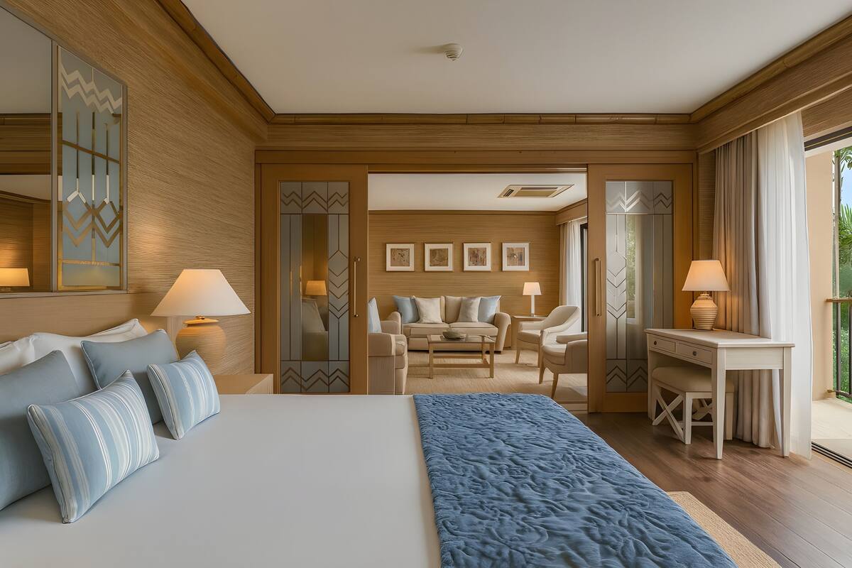Quarto de hotel com uma cama grande, uma mesa de escritório, um sofá e uma mesa de jantar. Há luminárias em ambos os lados do quarto e uma janela grande à direita.