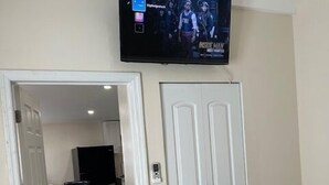 A room with a television mounted above a white door, a dining area with a table and chairs, and a bed with a plaid blanket. - Charming secure 2 bedroom home near Miami AirPort. Free parking (Miami)
