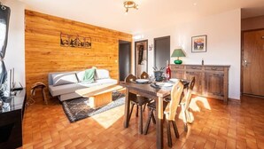 A cozy living room with a wooden wall, a sofa, a dining table with chairs, and a kitchen area with cabinets. - Le p'tit Schuss (La Giettaz)