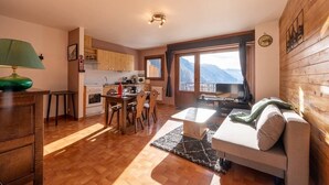 A cozy living room with a sofa, a dining table, and chairs. There is a kitchen area with wooden cabinets and a stove. A large window offers a view of mountains. - Le p'tit Schuss (La Giettaz)
