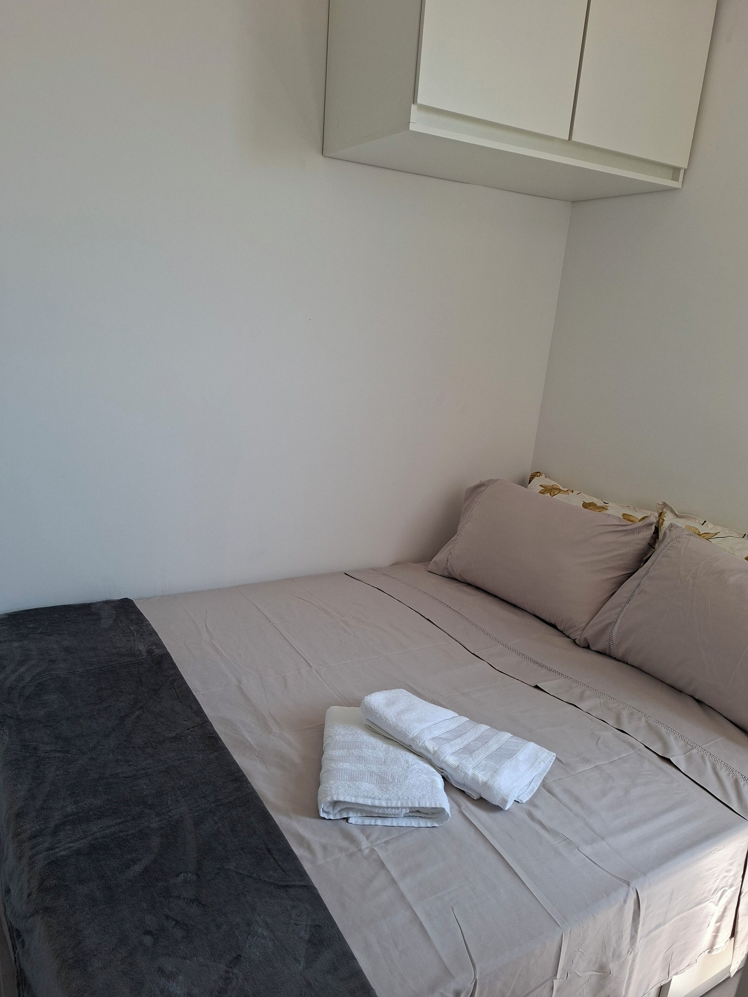 A neatly made bed with beige bedding and a dark grey blanket, a white pillow, and two folded white towels on the bed.