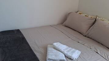 A neatly made bed with beige bedding and a dark grey blanket, a white pillow, and two folded white towels on the bed.
