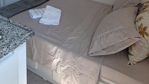 Een netjes opgemaakt bed met een beige sprei en kussens, een afstandsbediening en een flatscreen televisie aan de muur.
