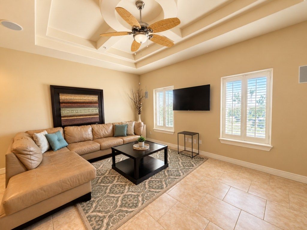 A living room with a sectional sofa, a coffee table, a ceiling fan, a flat-screen TV mounted on the wall, and a patterned rug.