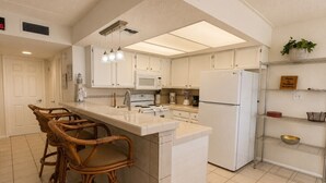 A kitchen with white cabinets, a white refrigerator, a microwave, a stove, and a sink. There are three wicker chairs and a white countertop. - Two Bedroom One Bath Off The Beach (South Padre Island)