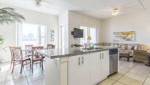 A modern kitchen with white cabinets, a granite countertop, and a built-in dishwasher. There is a dining area with wooden chairs and a table. A television is mounted on the wall, and a painting is visible in the living room. - Two Bedroom Two Bath Bayside Condo (South Padre Island)