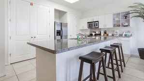 A modern kitchen with white cabinets, a granite countertop, a stainless steel refrigerator, a microwave, and a sink. There are three wooden bar stools and a potted plant. - Two Bedroom Two Bath Bayside Condo (South Padre Island)