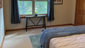 Ein Schlafzimmer mit einem Bett, einem fensterbelaubten Fenster, einem Schreibtisch mit Pflanze und einer Tür.