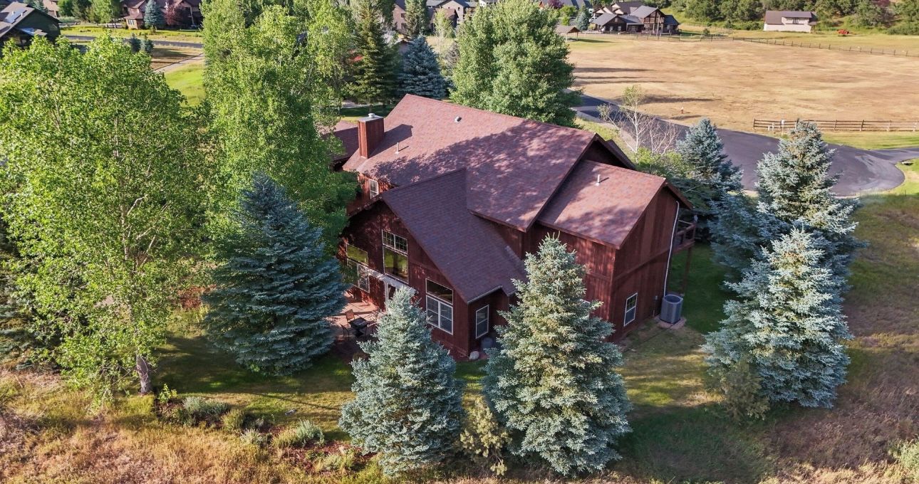 A large, single-story house with a red roof, surrounded by trees and a fenced yard.