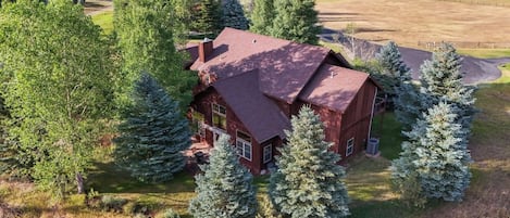 A large, single-story house with a red roof, surrounded by trees and a fenced yard.