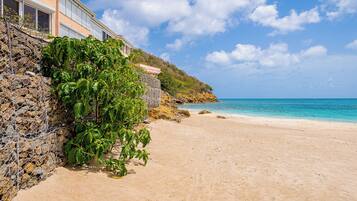A beach with a stone wall and vegetation, a clear blue sky, and a calm sea.