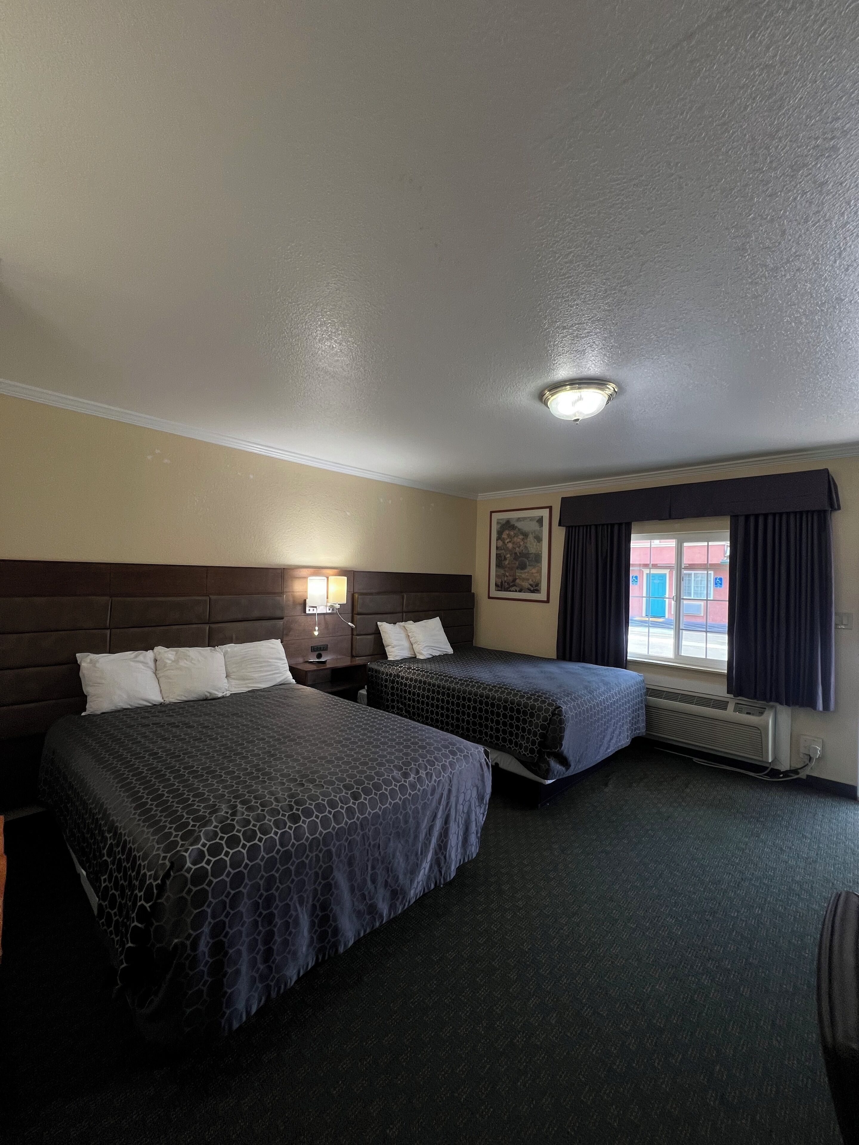 A hotel room with two beds, a nightstand, a lamp, a window with curtains, and a ceiling light.