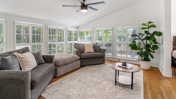 A spacious living room with a gray sectional sofa, a gray armchair, and a round coffee table. There is a ceiling fan, large windows with blinds, and a potted plant.
