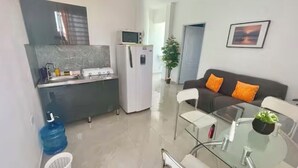 Eine moderne Küche mit Kühlschrank, Mikrowelle und Spüle. Es gibt einen Essbereich mit einem Glastisch und Stühlen. Im Wohnbereich befindet sich eine Couch mit orangenen Kissen.
