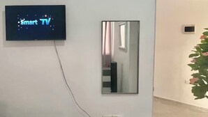 Ein Zimmer mit einem Bett, einem an der Wand montierten Fernseher mit der Aufschrift 'Smart TV', einem Spiegel und einer Topfpflanze.