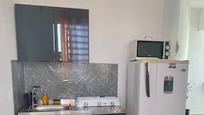 Eine kompakte Küche mit Gasherd, Mikrowelle und Kühlschrank. Es gibt einen Wasserspender und ein Waschbecken. Ein großer, blauer Wasserspeicher steht auf dem Boden.