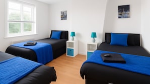 A hotel room with two beds, each with a blue bedspread and black headboards. There are two white nightstands with teal lamps, and a white shelf unit between the beds.