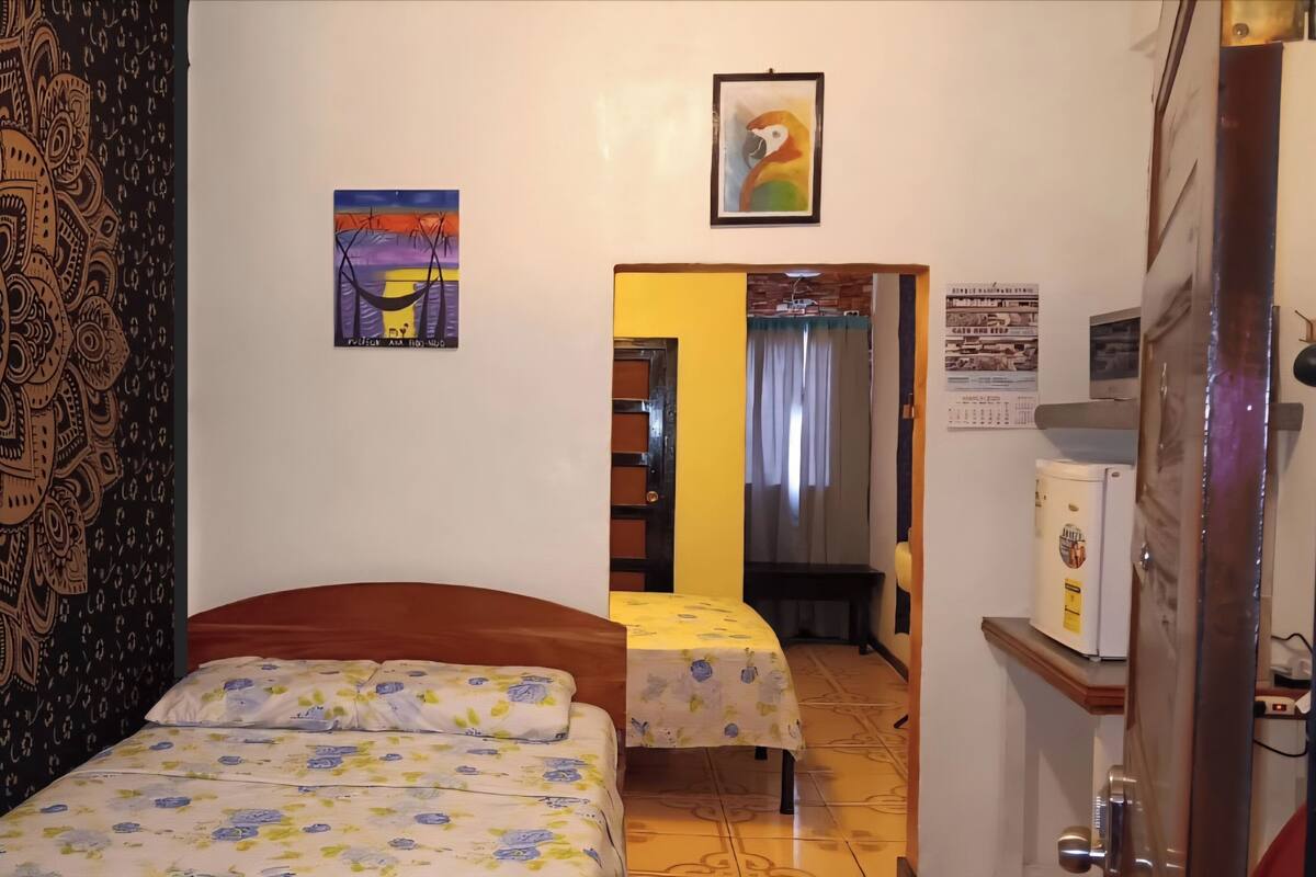 A room with a bed, a small table, a chair, a painting, and a door leading to another room.