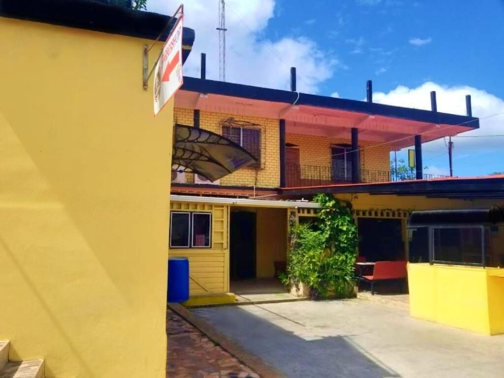 Ein gelbes Gebäude mit roter Markise, einem kleinen Garten und einer blauen Mülltonne.