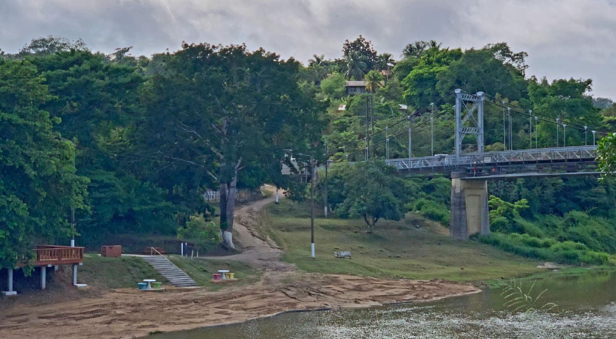 A suspension bridge over a river, surrounded by dense greenery and a small building with steps leading down to the water.