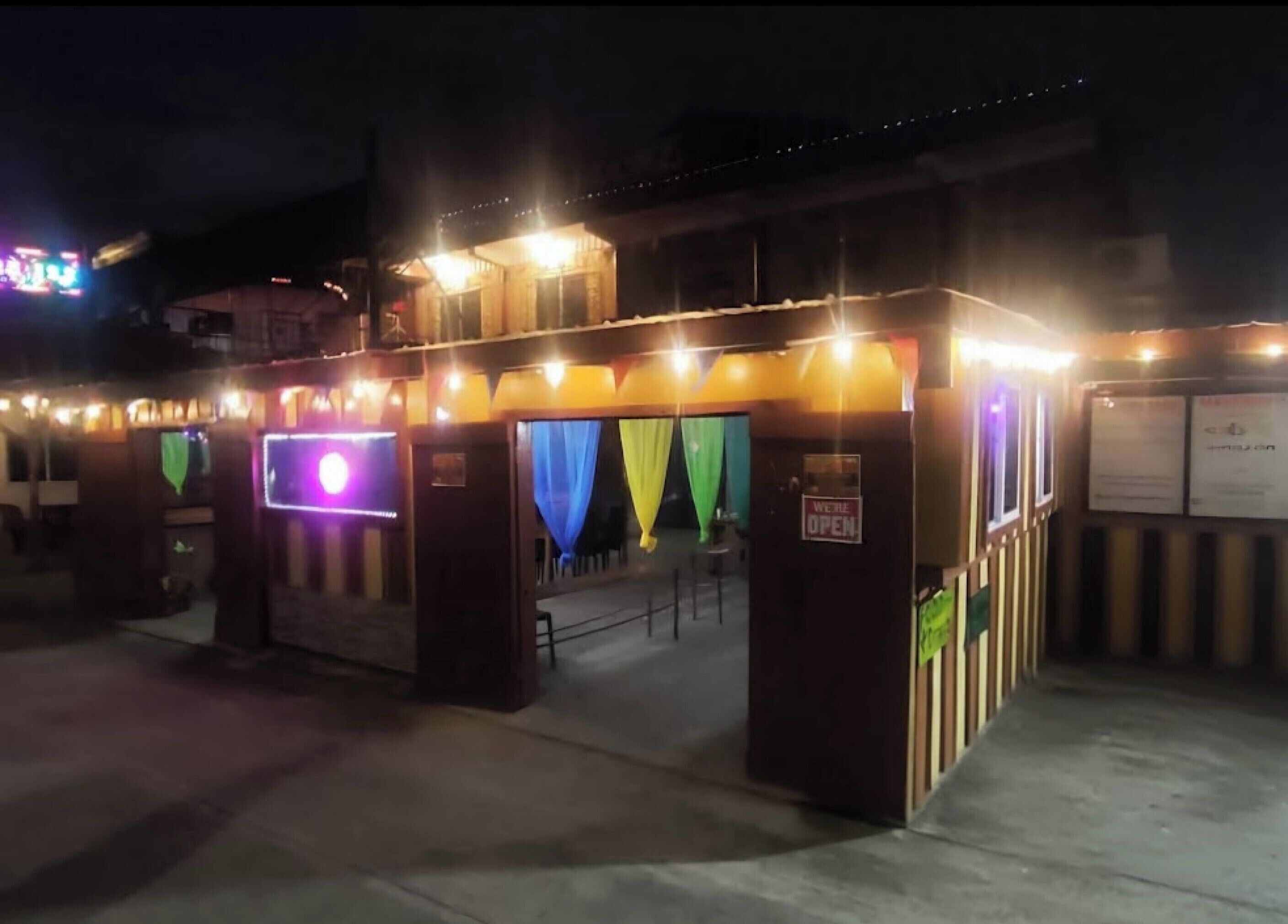 A building entrance with a 'WE ARE OPEN' sign, a covered area with colorful curtains, and a small seating area with a table and chairs.