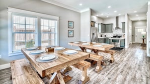 A spacious dining area with a large wooden table and benches, a kitchen in the background, and large windows with blinds. - exhale: Private heated pool, Free golf cart, 2 mins to beach, Bunk Rooms (Miramar Beach)