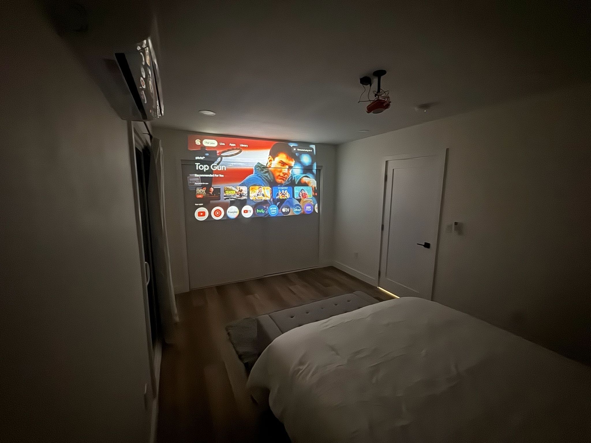 Un dormitorio con cama, proyector de pared que muestra un canal de televisión y ventilador de techo.