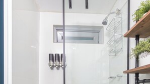 A modern bathroom with a glass-enclosed shower, a wooden stool, a patterned floor, and a shelf with plants.