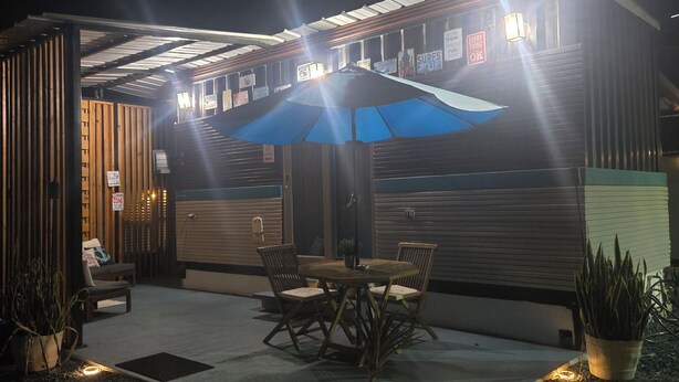 An outdoor seating area with a table and chairs under a blue umbrella, surrounded by potted plants and illuminated by ground lights.