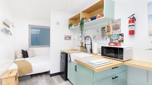 A compact kitchen with a microwave, sink, and a small refrigerator. There is a window above the bed and a shelf with various items.
